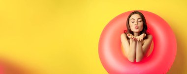 Portrait of beautiful female model posing with pink swimming beach ring, sending air kiss, mwah flirty, standing over yellow background.