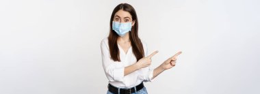 Portrait of corporate woman in face medical mask, pointing fingers left, showing logo, company banner, standing against white background.
