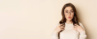 Close up portrait of beautiful girl in glasses, looking amazed and surprised, gasping excited, standing over beige background.