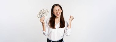 Joyful woman dancing with money, smiling pleased, winning prize, standing over white background.