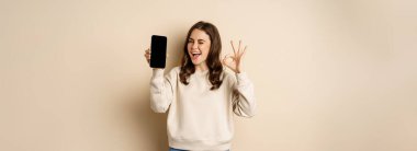 Beautiful modern woman winks, shows okay sign and mobile phone screen, app interfce, standing over beige background.