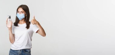 Covid-19, sağlık ve sosyal uzaklık kavramı. Tıbbi maske takmış, baş parmak ve el dezenfektanı gösteren mutlu, genç bir kızın portresi..