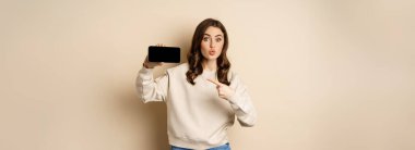 Image of girl pointing finger, showing mobile phone horizontal screen and looking amazed by application, standing over beige background.