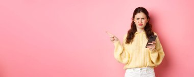 Hoşnutsuz kız somurtuyor, surat asıyor ve akıllı telefonu tutarken sol telif alanını işaret ediyor, pembe arka planda duruyor..