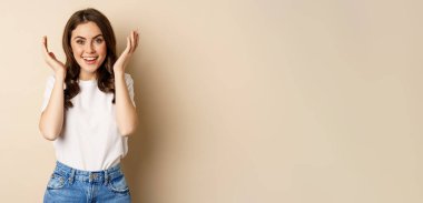 Surprise. Happy young woman open eyes with hands and waiting for gift, standing over beige background. Copy space
