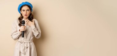 Image of insecure young woman standing with mic microphone and looking shocked worried, fear of speaking in public, standing over beige background.