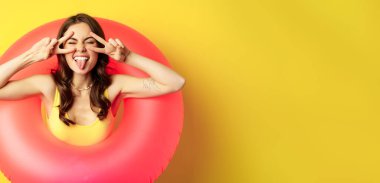 Portrait of attractive modern girl inside trendy pink beach swimming ring, showing peace v-sign and smiling happy, standing over yellow background.