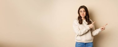 Skeptical girl looking hesitant, pointing fingers right at logo, promo text, standing over beige background.