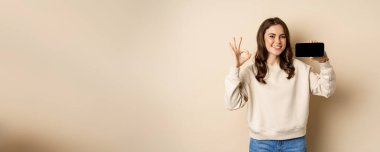 Smiling happy girl showing smartphone screen app, mobile interface, okay sign, standing over beige background.