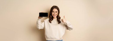 Smiling happy woman showing smartphone horizontal screen, thumbs up, recommending website, online store or app, standing over beige background.