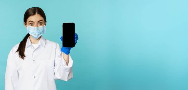 Young woman doctor in medical face mask and hospital uniform, showing mobile phone app, screen interface, online clinic concept, standing over torquoise background.