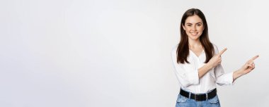 Portrait of smiling confident woman, business girl showing advertisement, pointing fingers right at logo, banner or announcement, standing over white background.