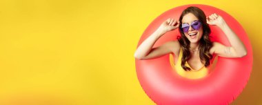 Close up portrait of stylish young woman, girl with swimming ring and summer sunglasses, ready for beach holiday, vacation, standing over yellow background.
