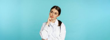 Thoughtful medical worker, young doctor woman looking unamused, bored, standing in white coat against blue background.