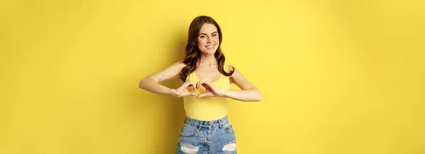 心を示す美しい若い女性 愛のサイン ジェスチャーのような 笑顔幸せとかわいいです 黄色の背景にタンクトップとショートパンツに立って — ストック写真