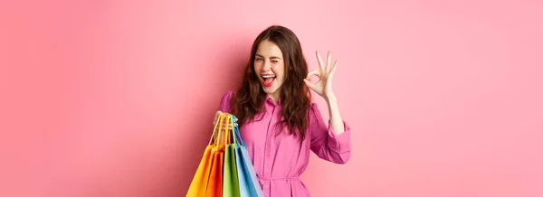 stock image Young happy woman shopper showing okay sign, winking pleased with good discounts, buying staff on sale, holding shopping bags and smiling pleased, pink background.