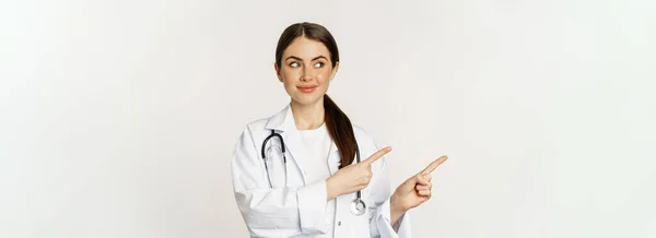笑顔の若い女性医師の肖像画 医療従事者 左指を指す クリニックのプロモーションを示す ロゴやバナー 白い背景の上に立って — ストック写真