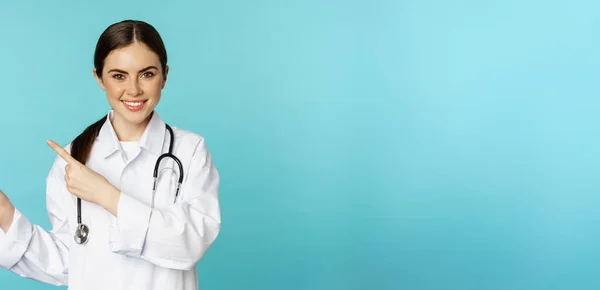 笑顔の医療従事者の肖像 聴診器付き白衣の少女医師 指差し指左 医療クリニックの広告を表示 ターコイズの背景 — ストック写真