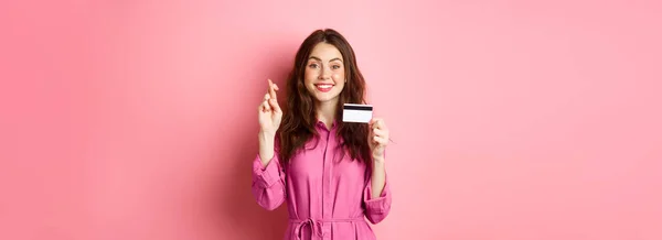 stock image Image of hopeful young woman showing plastic credit card and cross fingers for good luck, making wish and smiling, standing against pink background.