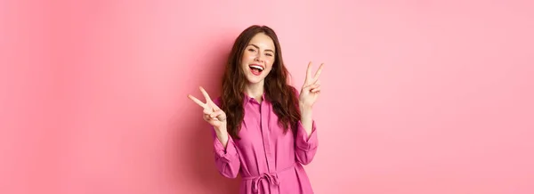 ポジティブな美しい女の子でスタイリッシュなドレス ショーVサイン平和と笑顔幸せな ポーズ近くピンクの背景 — ストック写真