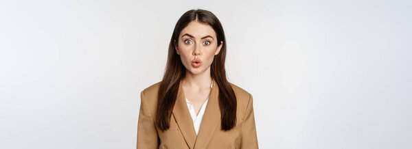 Portrait of office lady at workplace, looking surprised and curious, wow face, standing over white background in brown suit.