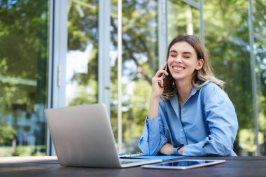 Ofis binasının yakınındaki temiz havada çalışan satış elemanının portresi. Bilgisayarlı gülümseyen şirket kadını, telefon görüşmesi yapıyor..