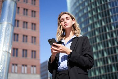 Genç bayan yöneticinin düşük açılı görüntüsü, takım elbiseli iş kadını, elinde cep telefonu, elinde akıllı telefonuyla caddede dikilirken..