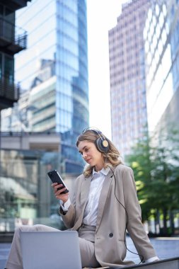 Takım elbiseli bir kadın, şehirde oturuyor, dizüstü bilgisayarla müzik dinliyor, cep telefonundan haber alıyor, yemek molasında dinleniyor..