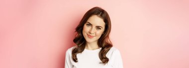 Close up of enthusiastic brunette girl with makeup, smiling and looking happy at camera, posing against pink background.
