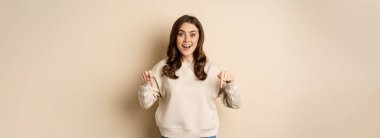 Enthusiastic attractive girl pointing fingers down, showing advertisement, link or promo below, standing over beige background.