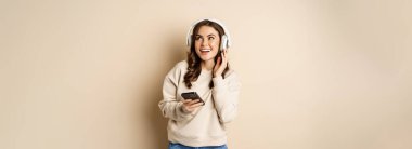 Happy caucasian woman listening music in headphones on smartphone app, dancing and having fun, standing over beige background.