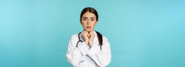 Worried and concerned physician, hospital worker in white lab coat, looking scared and nervous at camera, standing with stethoscope over torquoise background.
