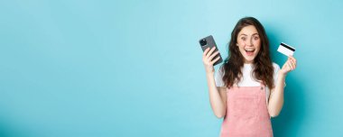 Neşeli kız kameraya heyecanlı görünüyor, akıllı telefon ve plastik kredi kartı tutuyor, mavi arka planda duruyor..