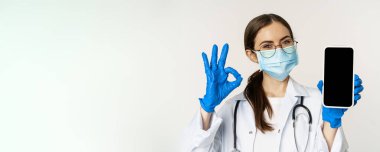 Online medical help concept. Enthusiastic young woman doctor in face mask, showing okay sign and mobile phone app, smartphone screen interface, white background.