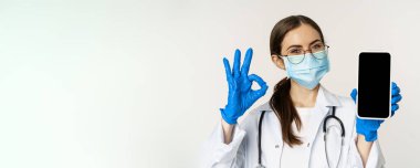 Çevrimiçi tıbbi yardım konsepti. Hevesli maskeli genç bayan doktor, iyi bir işaret ve cep telefonu uygulaması gösteriyor, akıllı telefon ekranı arayüzü, beyaz arka plan.