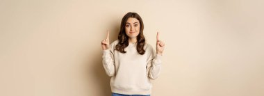 Smiling young woman looking indecisive, shrugging and smirking while pointing fingers up, standing over beige background.