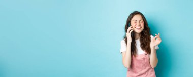 Güzel ve şık bir kızın portresi telefonla konuşurken gülüyor, akıllı telefonu kulağının dibinde tutuyor ve gülümsüyor, sohbet ediyor, mavi arka planda dikiliyor..