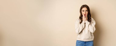 Portrait of shocked brunette woman drop jaw, gasping and staring speechless at camera, beige background. Copy space