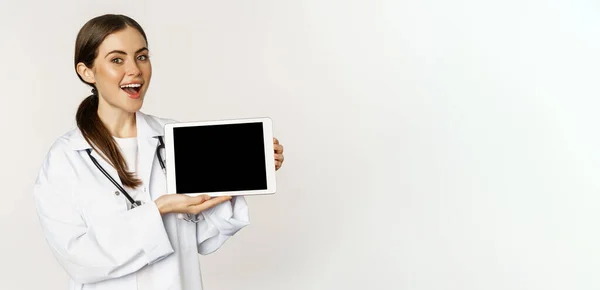 stock image Online medical help, remote appointment. Smiling beautiful woman doctor showing digital tablet screen, demonstrating website logo, standing over white background.