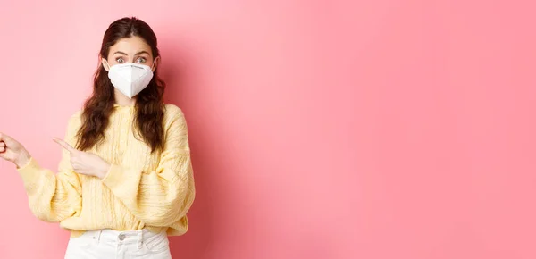 Covid 封锁和大流行病概念 年轻而好奇的女性展示了促销文稿 把手指放在一边 戴着呼吸口罩以采取预防措施 粉红的背景 — 图库照片