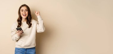 Happy brunette girl holding smartphone and cheering, winning, celebrating win on mobile phone app, standing over beige background.