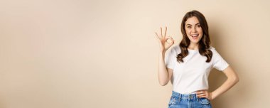 Beautiful female model smiling pleased, showing okay, zero sign, complimenting, praising you, standing over beige background.