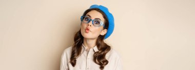 Young funny woman acting, fooling around, pucker lips and grimacing, posing against beige background in trendy blue hat.