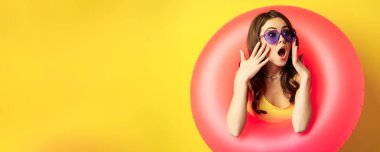 Portrait of young pretty woman in sunglasses, wearing beach swimming ring, looking surprised and amazed, shocked wow face, yellow background.