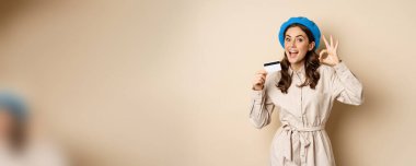 Concept of shopping and sale. Happy young woman in stylish clothes, showing credit card store discount and okay, satisfied, like and approve smth, standing over beige background.