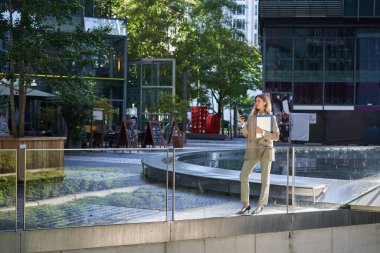 Takım elbiseli şık bir kadın, dizüstü bilgisayar ve iş belgeleriyle ayakta duruyor, cep telefonu kullanıyor, akıllı telefon ekranına bakıyor..
