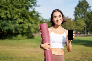 Heyecanlı fitness kızı spor ve egzersiz için başvuru öneriyor, telefon ekranını gösteriyor, antrenmandan sonra parkta kauçuk yoga minderiyle duruyor..