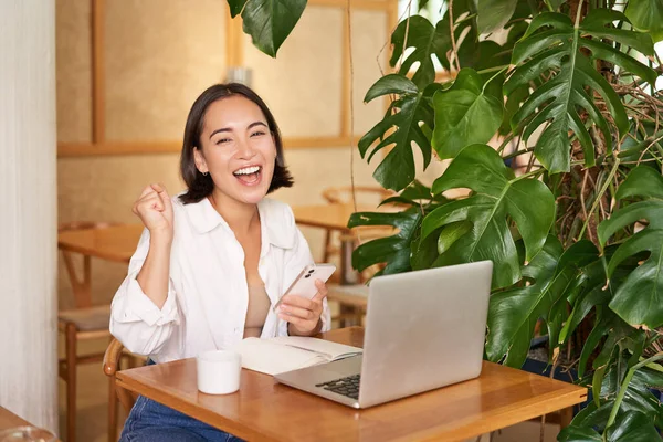 stock image Happy young asian woman feel success, winning and celebrating, sitting with laptop and smartphone in cafe and triumphing, shouting yes.