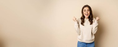 Enthusiastic brunette woman laughing and smiling, cheering, shaking hands and celebrating, triumphing, standing over beige background.