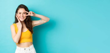 Temiz tenli ve mükemmel vücutlu güzel bir kadının portresi, yazlık kıyafetler giyiyor, barış işareti gösteriyor ve yüzlerine dokunuyor, mavi arka planda duruyor..